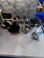 Cooking items arranged on a table including 2 metal pots with lids and black handles, 1 frying pan, 2 metal colanders/strainers, 2 glass baking dishes, 1 metal baking pan, and 1 glass measuring cup with blue handle.