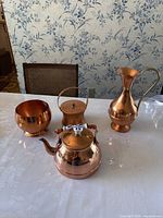 Set of four copper items on white tablecloth including bowl, storage container, tea pot, and water jug next to a chair and floral wallpaper background.