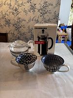 Bodum Chambord French press coffee maker in box, Pyrex glass teapot, and two ceramic mugs on table with floral wallpaper background