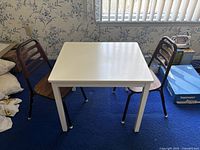 White rectangular kids table with two brown wooden and metal chairs placed on blue carpeting in a room with floral wallpaper.