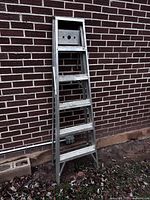 Metal A-frame ladder standing against a brick wall, showing all five steps and the top platform with holes.