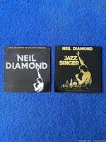 Two black album covers of Neil Diamond vinyl records '20 Golden Greats' and 'Jazz Singer' placed side by side on blue carpet.