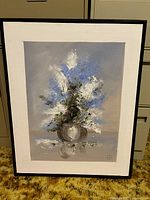 Framed painting displayed against a filing cabinet showing a vase with white and blue flowers on a gray background, signed lower right.