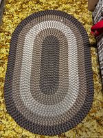 Full view of the oval braided rug laid flat on a yellow and brown patterned carpet, showing its detailed color bands and texture.
