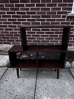 Front view of the retro wooden bookshelf showing open upper shelf and divided lower compartments with angled legs, positioned against brick wall.