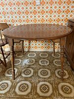 Round wooden dining table with turned legs, medium brown finish, against patterned wallpaper and tiled floor