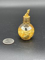 Front view of small round glass bottle with visible gold flakes inside, labeled as Genuine Gold Alaska. Cap has sculpted animal figure on top, placed next to a quarter for scale.