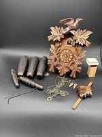 Full lot showing cuckoo clock body, four pine cone weights, clock chains, keys, pendulum and other small parts arranged on black background.