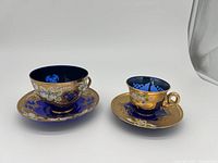 Photo of two hand-painted blue and gold teacups with matching saucers, viewed at an angle showing detailed decoration