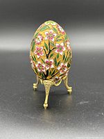 Frontal view of the cloisonné decorative egg on its gold-toned metal stand showcasing the intricate pink floral and green leaf enamel design on a gold background.