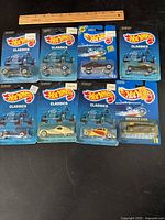 Top-down view of eight Hot Wheels Classics diecast cars in original packaging arranged on a black surface with a wooden ruler for scale.