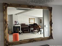 Large horizontal rectangular mirror with ornate gilded gold frame with intricate carvings reflected in room with piano and fireplace.