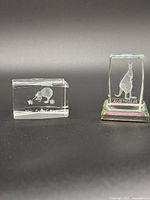 Two small clear glass paperweights side by side on dark surface with a kangaroo design marked 'AUSTRALIA' and a kiwi bird marked 'New Zealand'.