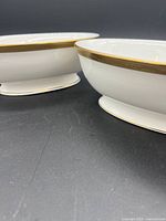 Side view showing two white oval footed vegetable bowls with gold trim around the edge.