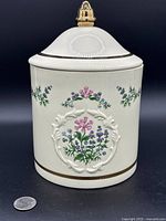 Side view of Lenox Spice Garden porcelain canister showing embossed floral pattern and gold rim detail with lid on top showing gold-accented finial.