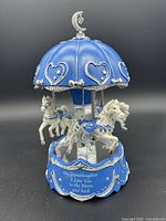 Front view of blue and silver musical carousel with three horses, crescent moon and dangling heart atop, and inscription 'My Granddaughter I Love You to the Moon and back'.