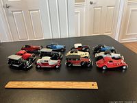 Overview of 12 assorted vintage model cars arranged on black table next to ruler for size reference