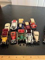 Top-down photo of 11 assorted miniature vintage cars in various colors including cream, red, yellow and green, all styled to resemble early 20th century automobiles on a black surface next to a ruler for size reference.