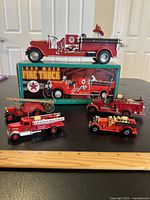 Five vintage red metal toy fire trucks displayed on a black surface, one is displayed on its box labeled '1929 Mack Fire Truck'. Various sizes and detailed features visible.