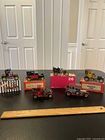 Photo showing all 6 vintage toy cars displayed with their corresponding branded boxes and a wooden ruler for scale on black table.