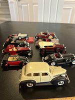 Nine assorted vintage metal toy cars arranged on a flat black surface, showing various angles and colors including cream, red, maroon, and black.