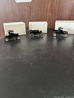 Front view of three black vintage diecast Ford Model T vehicles with white and gold spoked wheels arranged in front of their white boxes.