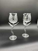 Pair of clear glass wine glasses shaped like roses with curved stems and leaf details on a black background.