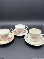 Front view of the three floral teacups and saucers arranged side by side on a black background, showing distinct floral patterns and gold trim details.