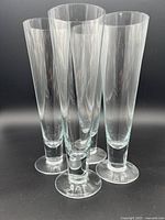 Set of four clear crystal pilsner glasses arranged close together, showing tapered cylindrical shape and solid footed base.