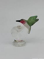 Front and side view of Swarovski miniature heart with hummingbird figurine showing green hummingbird with pink throat perched beside faceted heart containing small roses on clear crystal flower base.