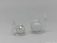Front view of two clear glass miniature snail figurines with faceted crystal shells reflecting rainbow colors, placed side-by-side on a white surface.