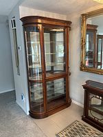 Front-side angle view of tall wooden curio cabinet with curved glass sides and front door, located near a wall and mirror in living space.