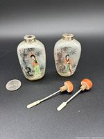 Two translucent glass snuff bottles with detailed hand-painted traditional Chinese designs of women and bamboo, each paired with a glass spoon with orange stopper. A quarter is included for scale.
