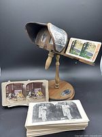 Wooden stereoviewer on stand with curved viewing hood, showing a stereograph card inserted for viewing along with additional cards displayed in front.