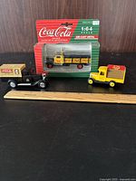 Three diecast Coca-Cola trucks displayed with a ruler for scale, one in original packaging and two loose models of vintage style trucks.