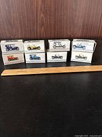 Eight original boxed vintage Readers Digest diecast cars arranged in two rows, showing the front and sides of the boxes with printed illustrations of different classic car models, accompanied by a 6 inch wooden artist's ruler for scale.