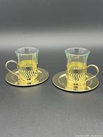 View of two vintage Schott Verran espresso cups with gold-tone metal sleeves and matching saucers, side by side.