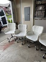 Four white vinyl rolling chairs arranged in a garage setting showing front and side angles, highlighting cushioned seats and backs with square pattern stitching.
