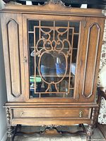 Front view of antique wooden china cabinet featuring glass door with decorative lattice woodwork and ornate carved details on top and drawer front.