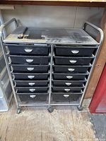 Full front view of black 14 drawer storage container with metal frame and wheels on wooden floor, drawers closed, showing two columns each with 7 drawers.