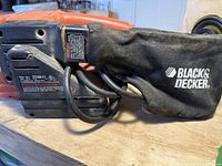 Side view of Black & Decker branded dust collection bag attached to belt sander showing power cord and labels.