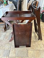 Two wooden butler trays, one standing on a folding wooden stand, the other leaning against the stand. Trays have built-in handles on the shorter sides.