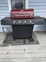 Front view of the red-hooded barbecue grill with four control knobs and side shelves, set on a wooden deck.