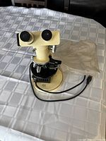 Front and eyepiece side view of Wild Heerbrugg microscope on a table showing binocular eyepieces, focusing knobs, stage, and power cord.