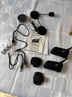 Top view of all items spread on white cloth showing two interphone devices, two wired headset earpieces, two USB cables, two mounting brackets, and user manual.