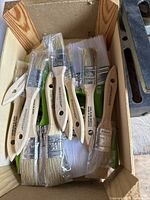 Box of natural bristle paint brushes with wooden handles, some wrapped in plastic.