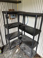 Two black heavy duty plastic shelving units each with 4 shelves, positioned side by side in a storage space with some dirt on and around the shelves.