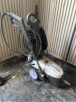 Front angled view of the Simoniz pressure washer with attached black hose and spray gun. Unit is on wooden floor inside a shed.