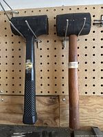 Two black mallets, one with a textured rubber handle and one with a wooden handle, hanging on a pegboard.