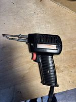 Front view of the black soldering tool showing dual metal tips, pistol grip, ventilation slits, and red trigger button on a wooden surface.